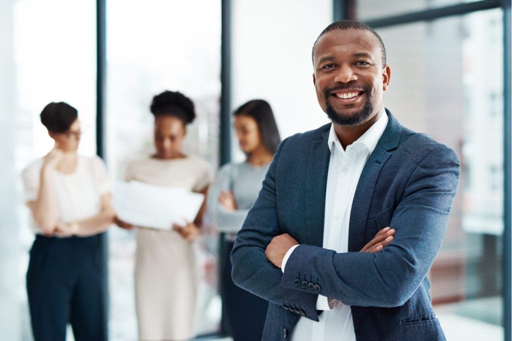 Confident businessman smiling with arms crossed, team in background.