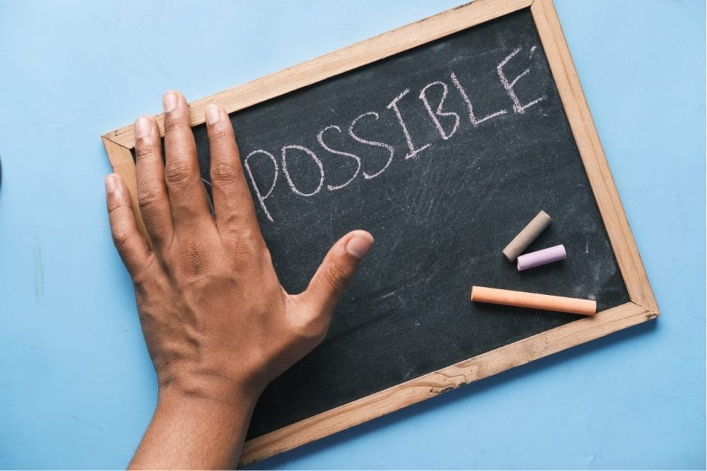 Hand rests on chalkboard displaying the word "POSSIBLE," alongside colorful chalk pieces.