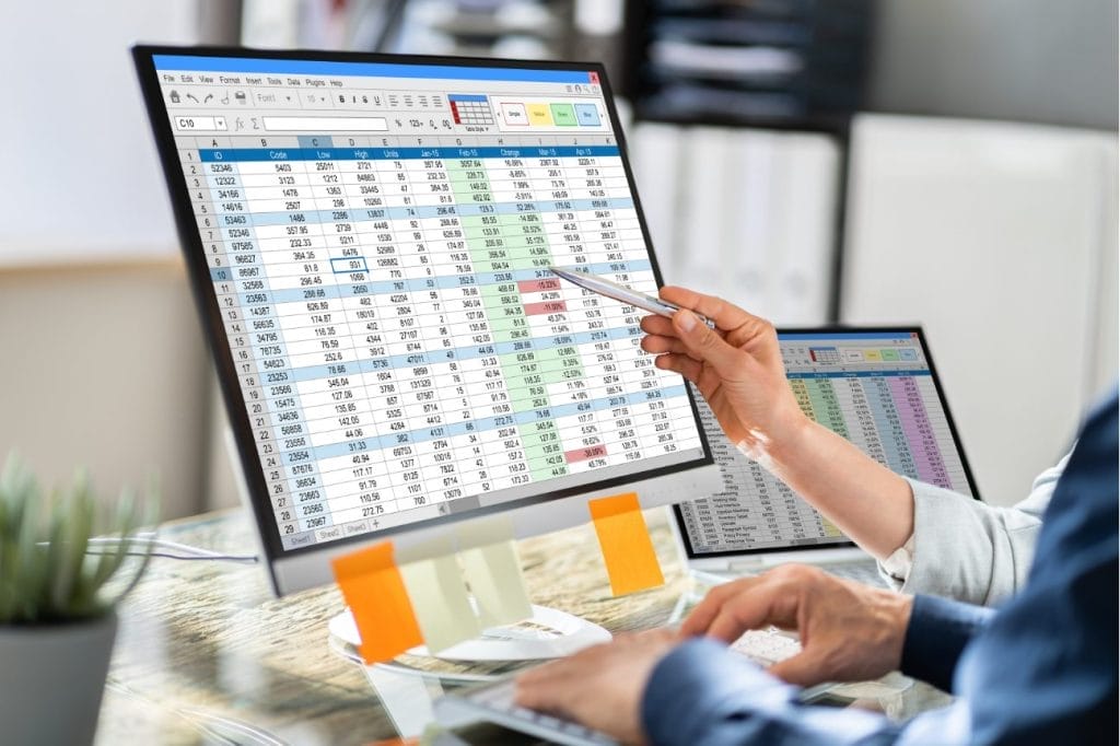 Two people analyzing financial data on computer screens. Spreadsheet shows sales figures.