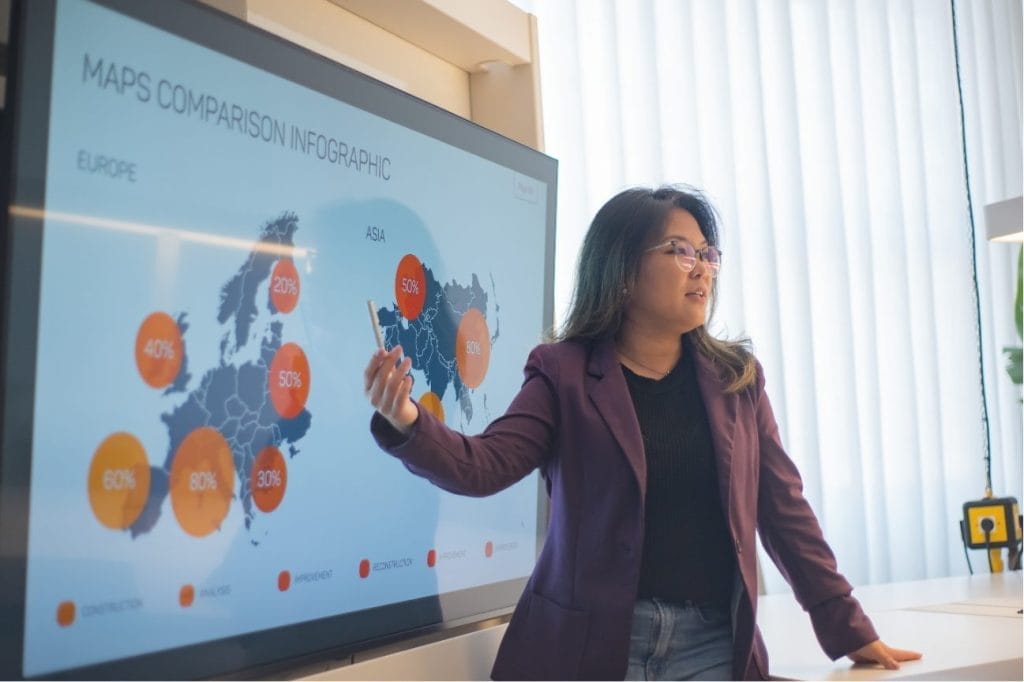 Woman presenting data infographic on interactive screen; maps comparison infographic shown.