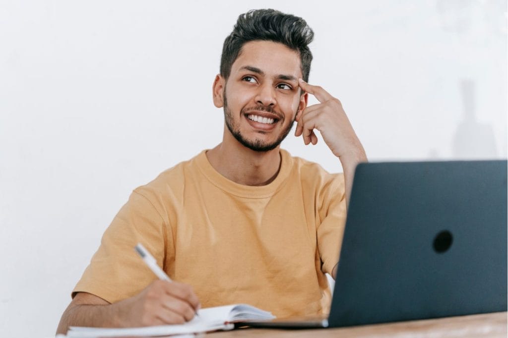 Happy male student taking notes while studying online.