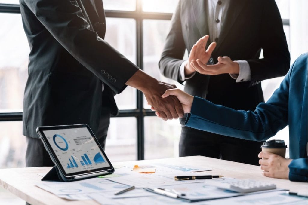 Businesspeople shaking hands, celebrating a successful deal. Financial charts displayed on tablet.