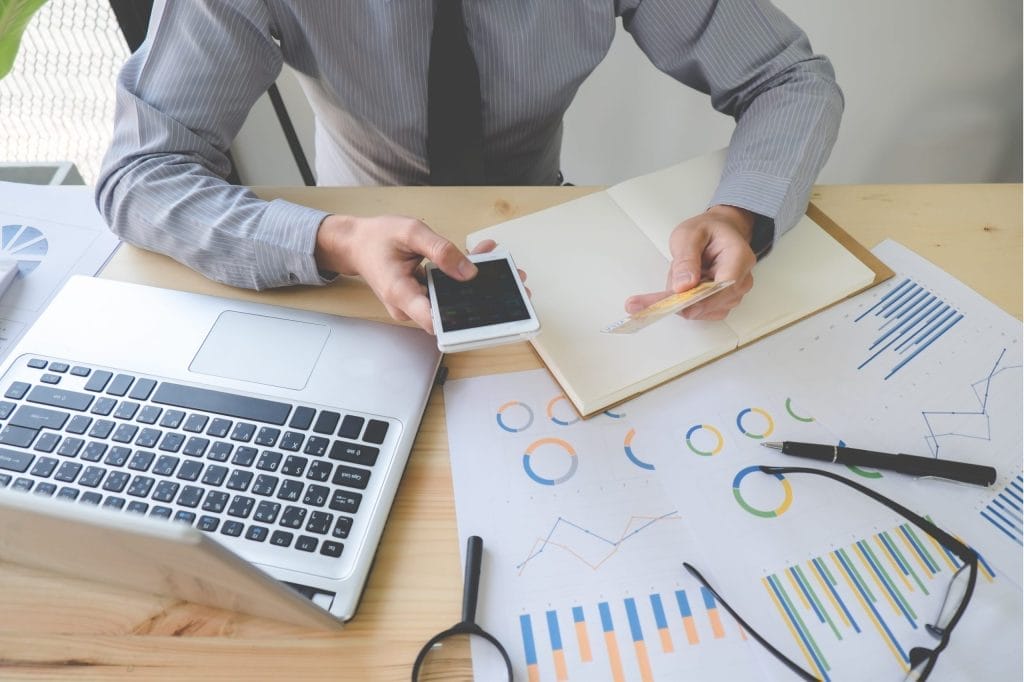 Businessman analyzes financial data with laptop, phone, graphs, and notebook on a wooden desk.